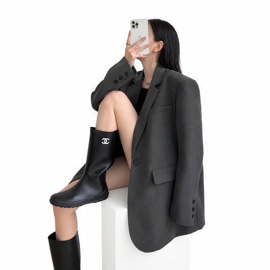 A woman takes a mirror selfie wearing an oversized dark gray blazer, a black top, and black leather rain boots with a silver interlocking CC logo on the side, seated on a white cylindrical pedestal against a plain light gray wall.